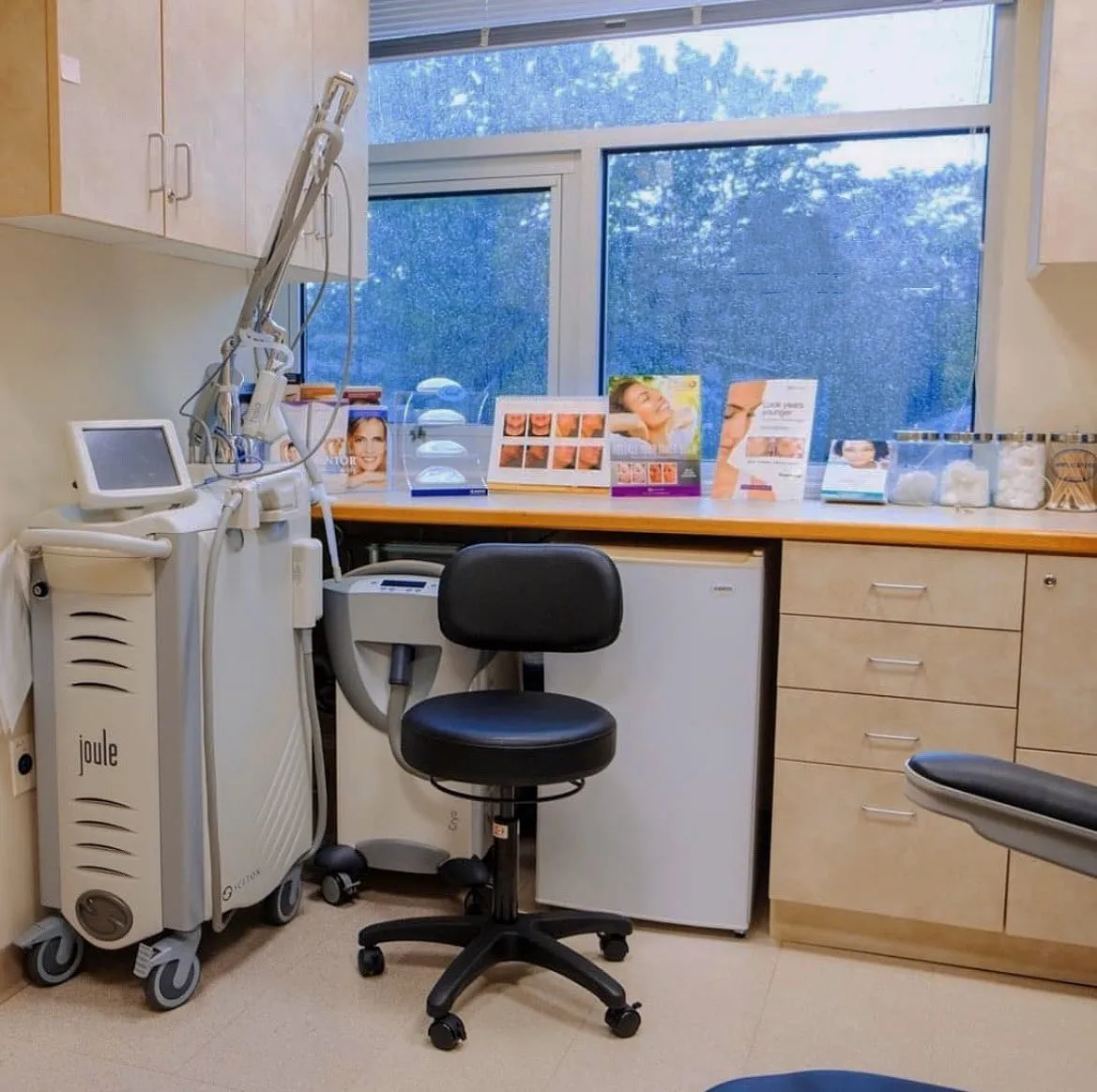 Non-surgical skin tightening treatment room with Joule device, consultation desk displaying before-and-after photos, and large windows overlooking a forested landscape